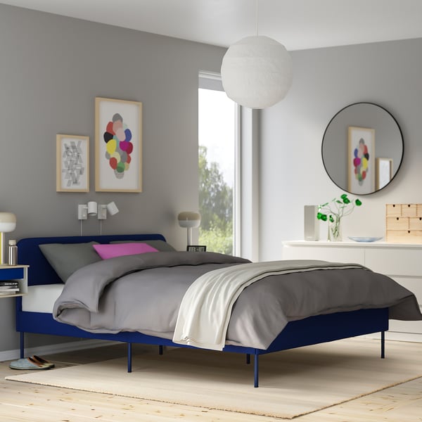 Bedroom with blue bed, grey blankets, colourful pillows, pendant light, off-white rug.