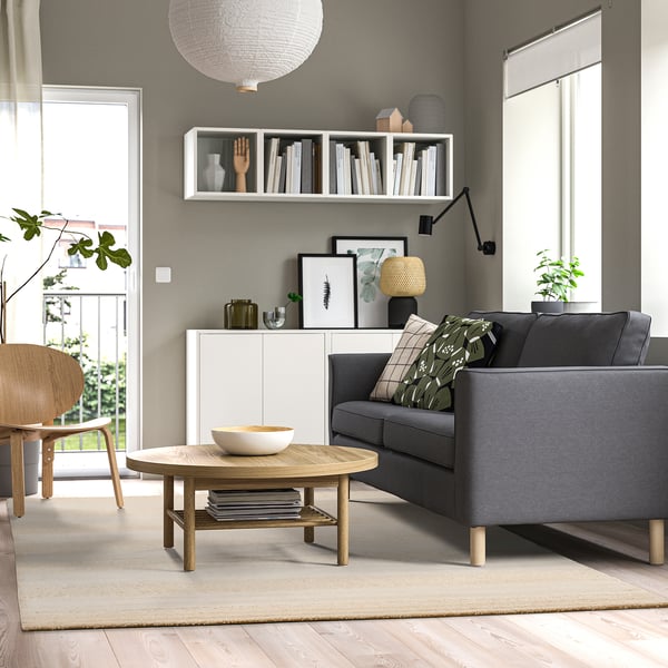 Modern living room with grey sofa, coffee table, rug. Two pillows with green patterns. Wall shelves with books and decor, wooden chair. Off-white rug.