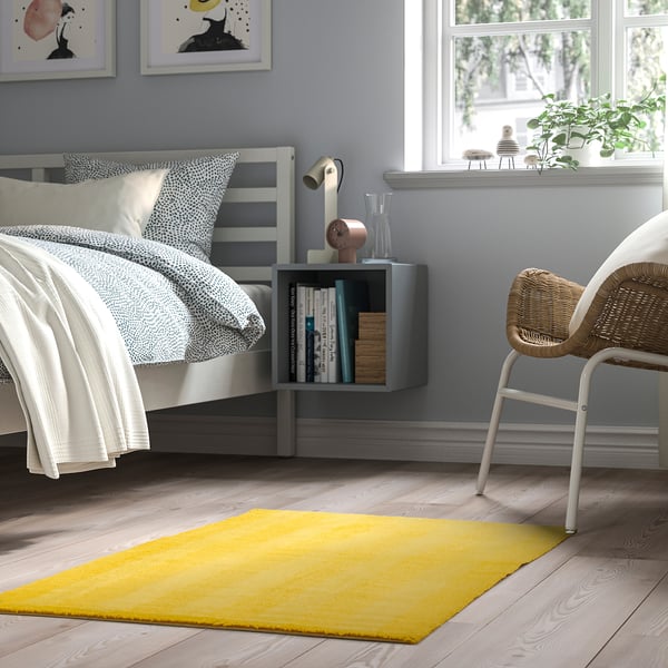 Bedroom with LANDSVÄG low pile yellow rug, small wicker chair, wooden floor, grey walls, and large bed. Bright rug contrasts the neutral room.