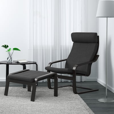 Modern, dark grey POÄNG armchair and footstool. Large, high-back design. Placed on a patterned rug beside a sleek side table with a plant.
