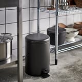 Small kitchen rubbish can, black with foot pedal, next to wire rack.