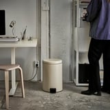 Person standing near a rubbish can and a desk with a computer and books.