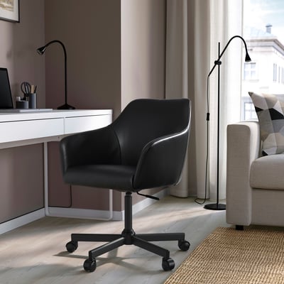 Modern home office: TOSSBERG black swivel chair at white desk with lamp, grey sofa and patterned pillow in background.