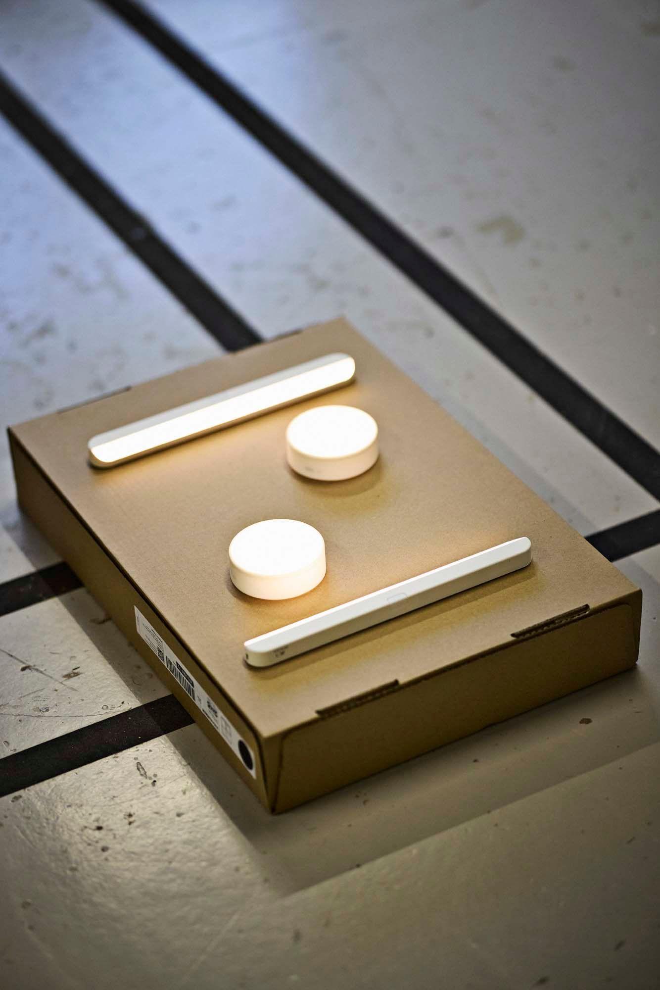 Two illuminated strip lights and two circular lights are placed on top of a cardboard flatpack package on a striped floor.