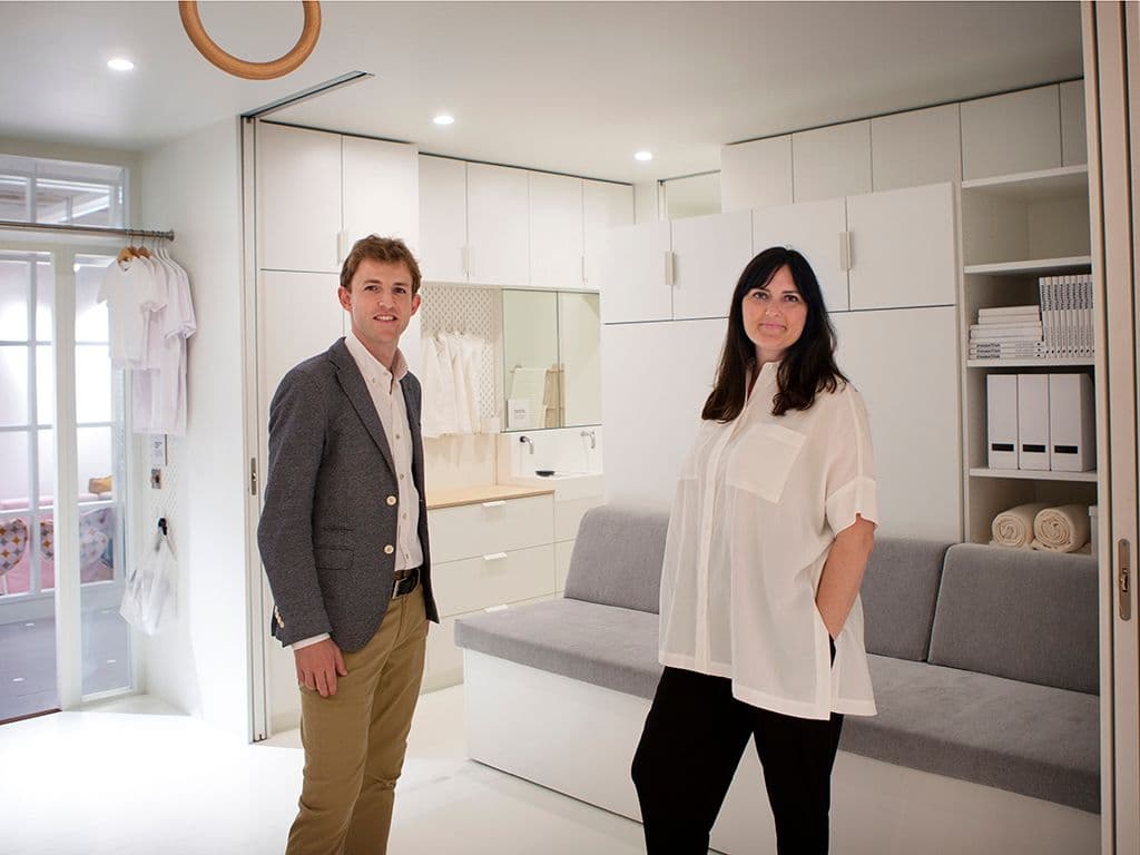 A man and a woman standing in a white room with a grey sofa and white storage behind them.