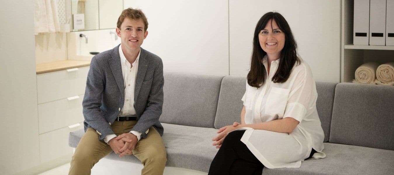 A man and a woman sitting on a grey sofa in front of white storage.