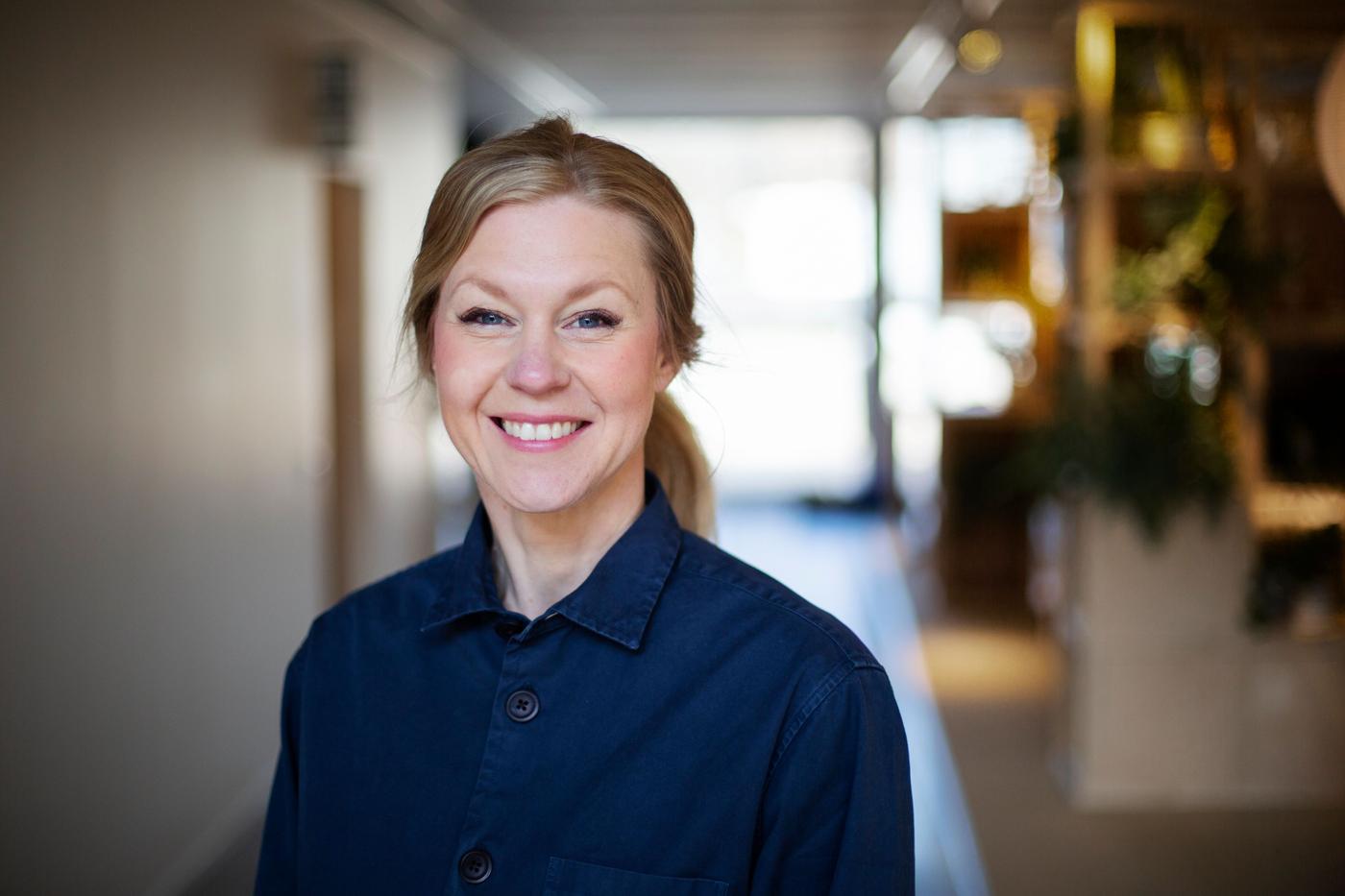 A woman with ligth hair and a navy blue shirt is standing in a blurred hallway and smiling towards the camera.