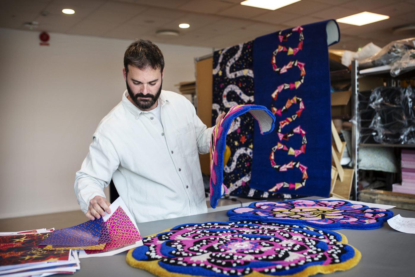 Luis Gomez Barquin looking at colourful printed patterns on a table beside two of his rugs with playful patterns.