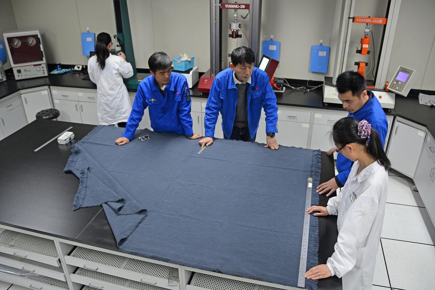 A group of people in blue and white work clothes measuring a blue rug on top of a workbench in a testing facility.
