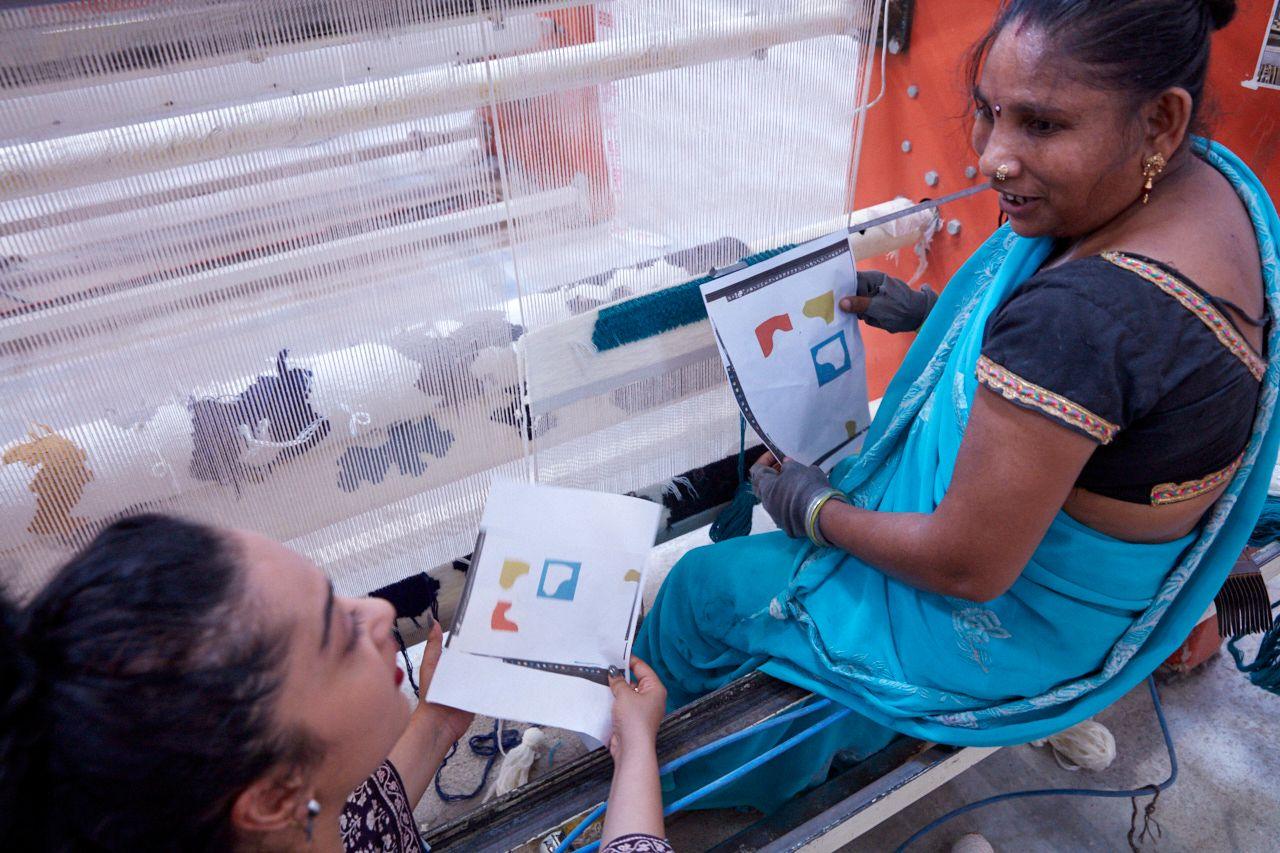 Designer Akanksha Deo and a woman sitting by a loom, looking at a multicolour pattern design on two pieces of paper.