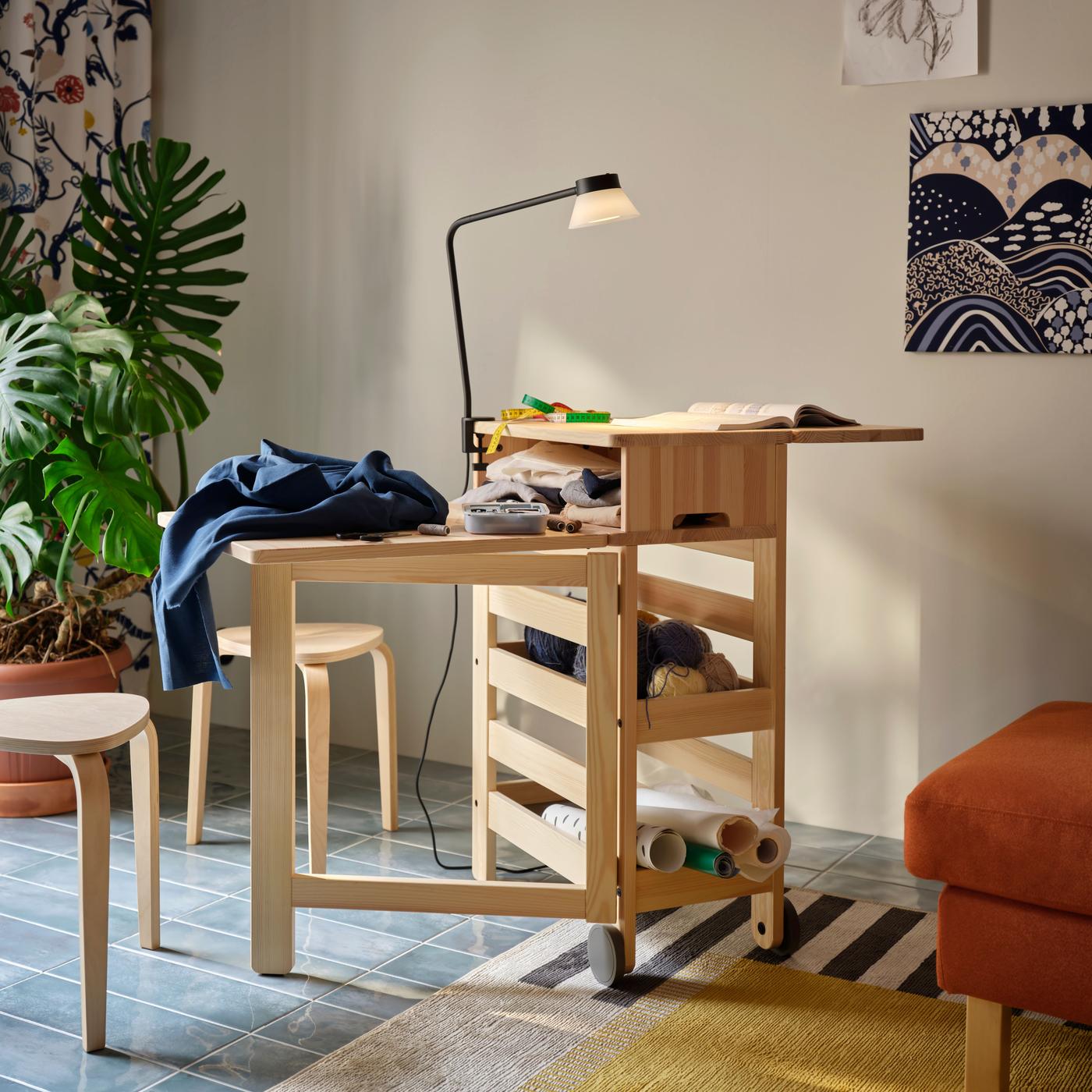 A living space with a wooden folding desk with shelves containing books and decor, alongside wood stools, an orange sofa and a leafy green plant.