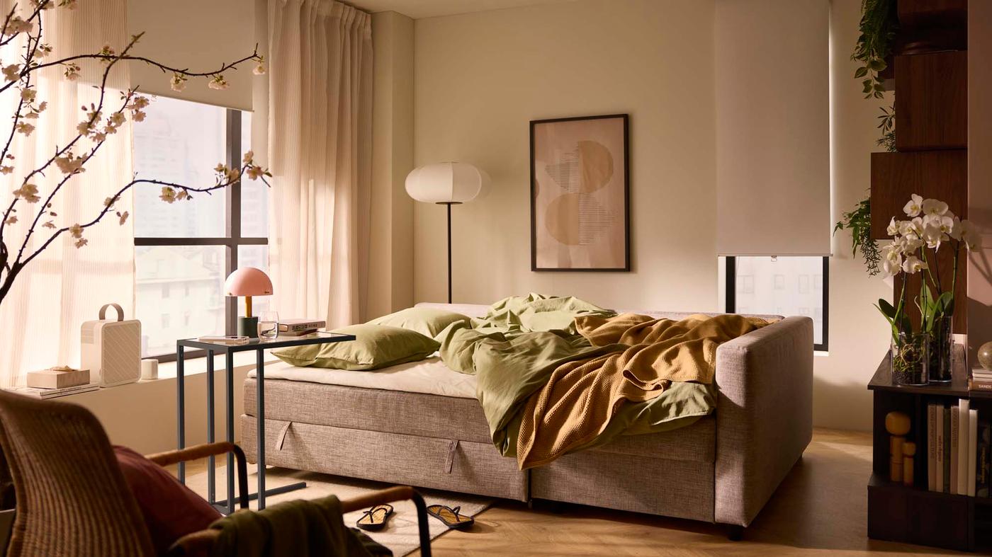 A warm space with tan sofa bed covered in green and brown bedding. There is a small metal table nearby with a lamp and other decor elements in the background.