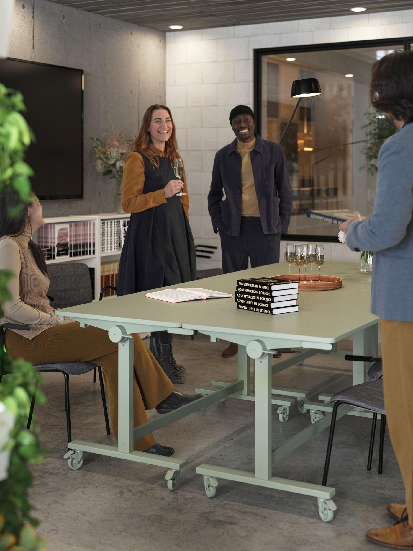 People in a meeting room, three standing, one sitting, all smiling, around two green desks that are placed next to each other.