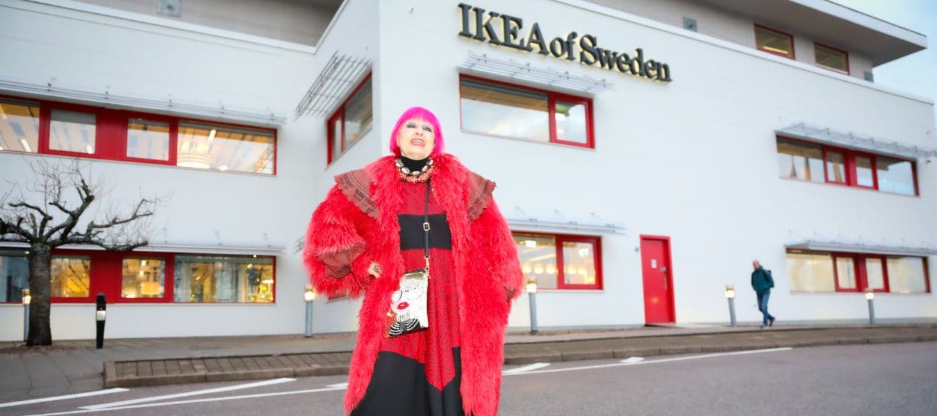 Designer Zandra Rhodes dressed in a red-black dress, red fake fur and accessories posing in front of the IKEA of Sweden building.