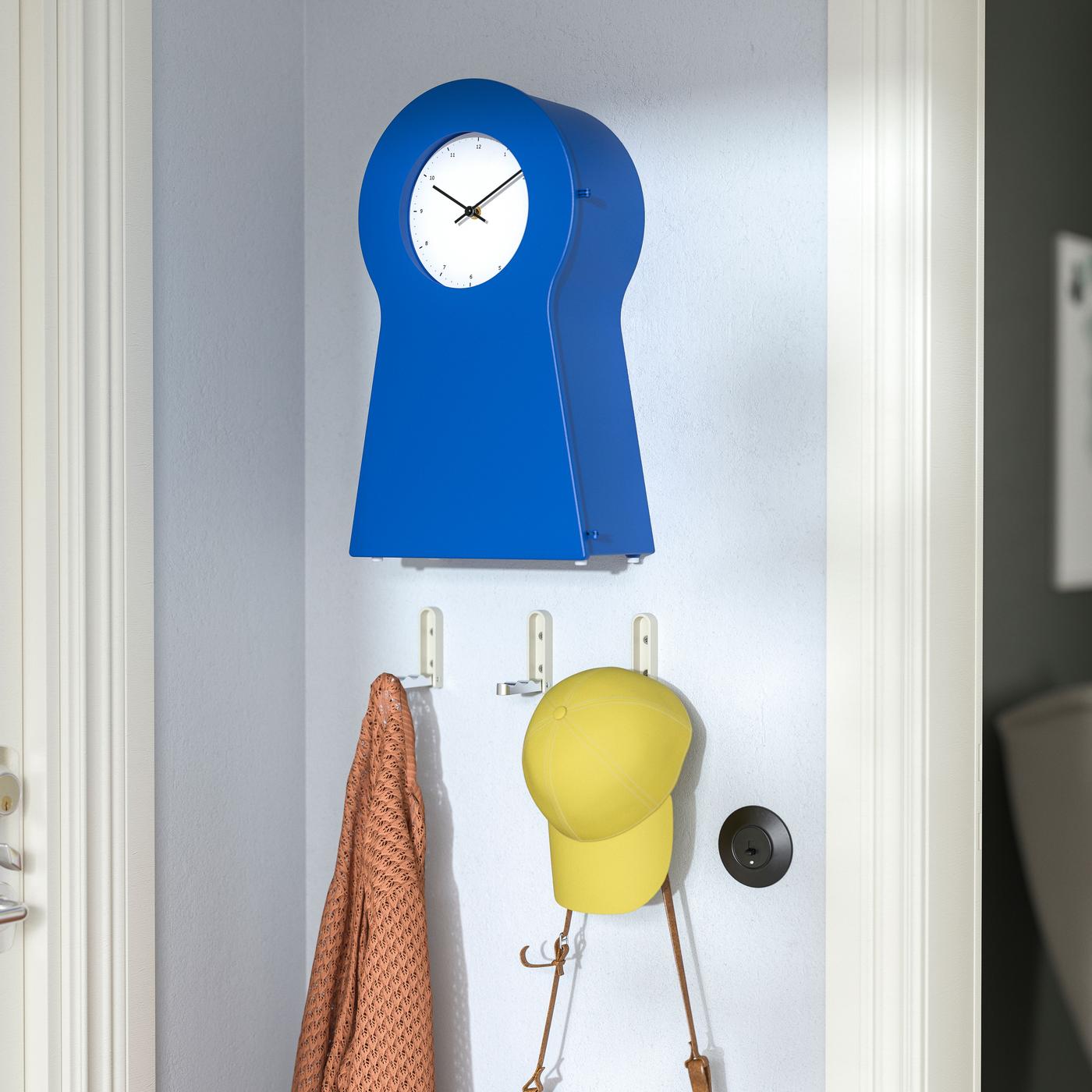 A blue clock on a wall above three hooks holding a yellow cap and an orange garment.
