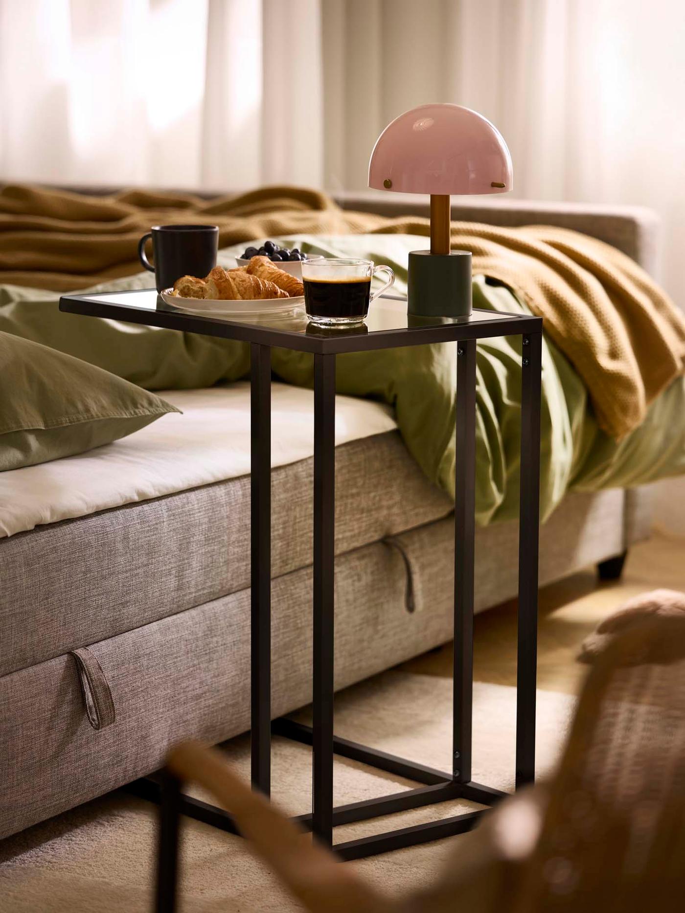 A small dark metal side table with frame partially tucked under a sofa, featuring a pink lamp and cups, with bedding on top of the sofa.