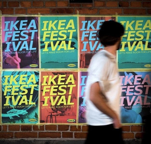 A person in a white top walks past a wall with 8 IKEA Festival posters taped on it.