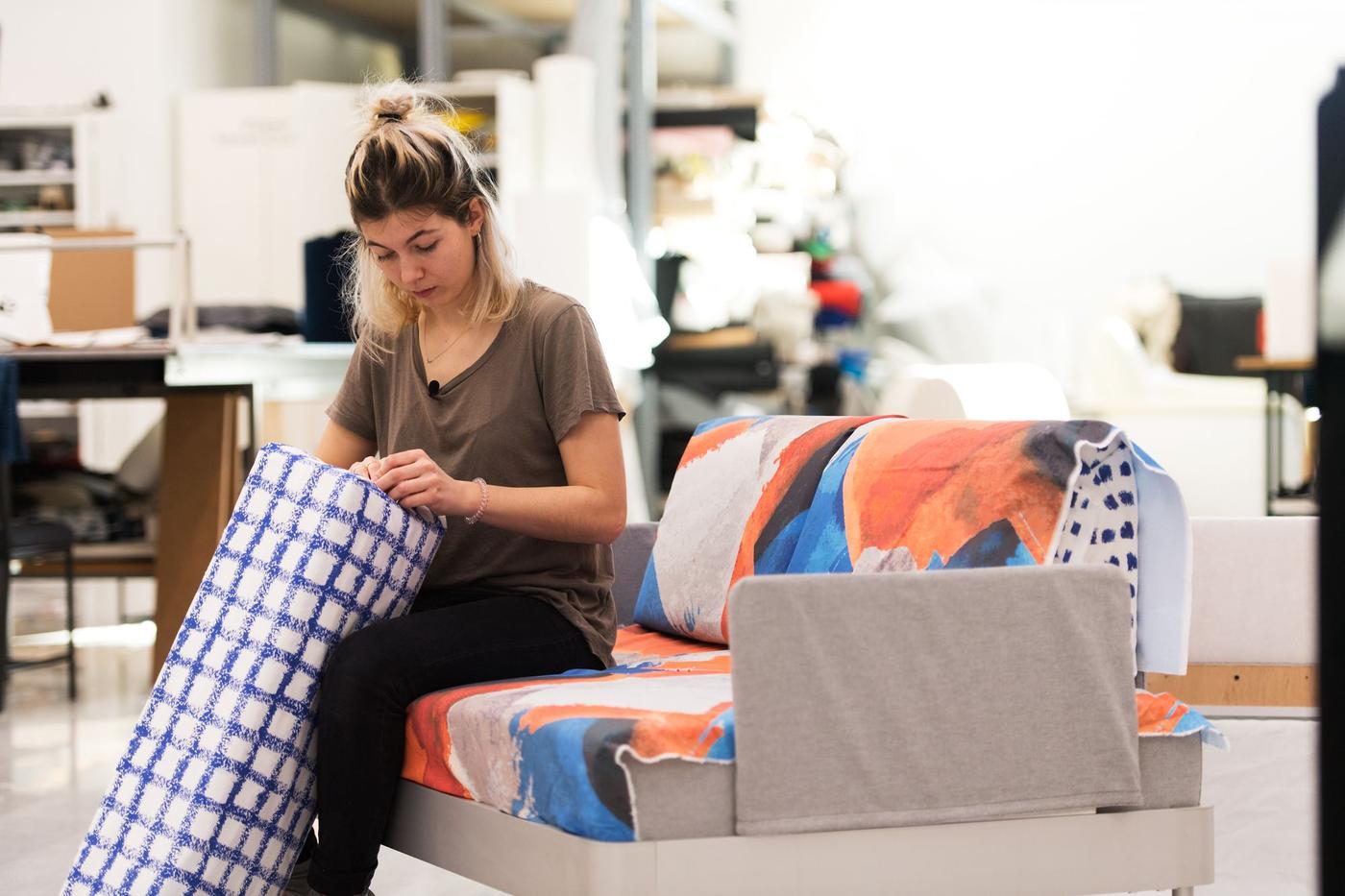 A woman sits on a coloured sofa, she is working with a blue and white check cushion.