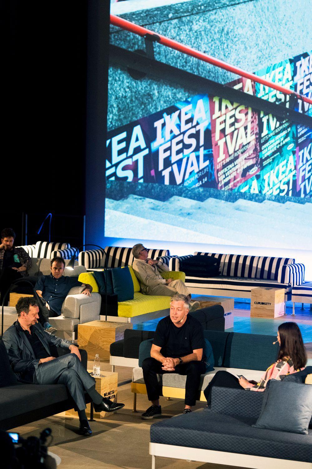 People sit on chairs and sofas in a big hall against a graphic backdrop.