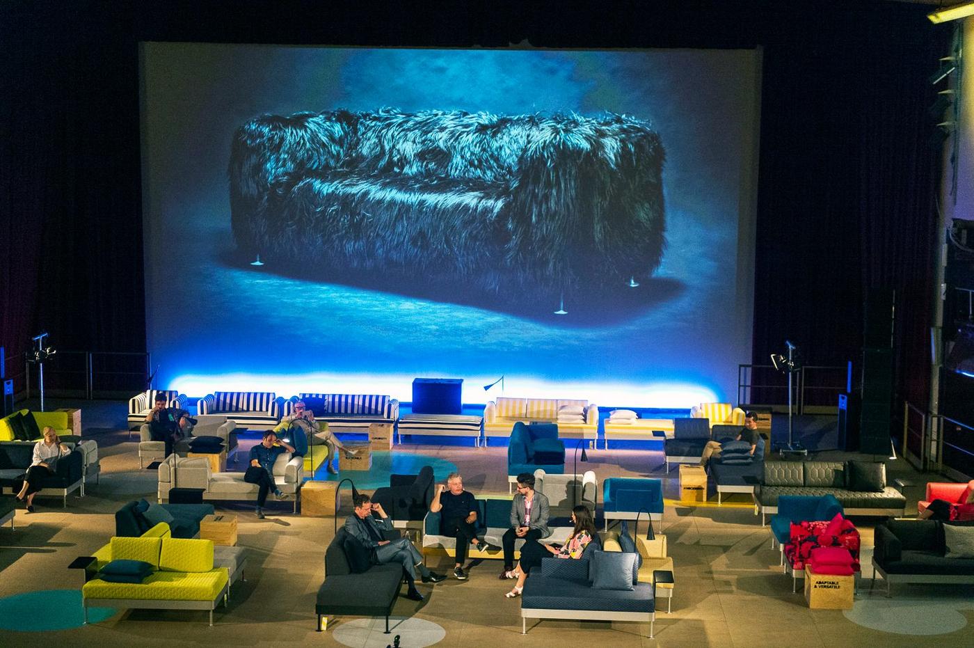 A large hall filled with chairs and sofas. In the centre a group of people talk. There is a giant photo of a sofa in the background.