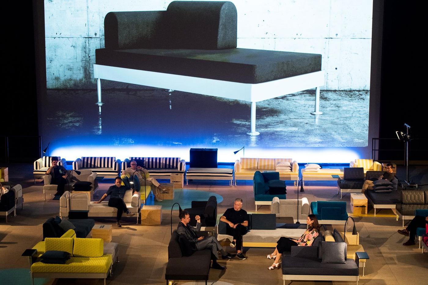 A large hall filled with chairs and sofas. In the centre a group of people talk. There is a giant photo of a chaise longue in the background.