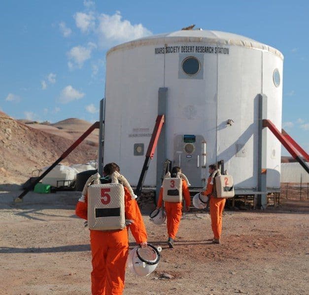 Three people in orange suits approach a large white structure.