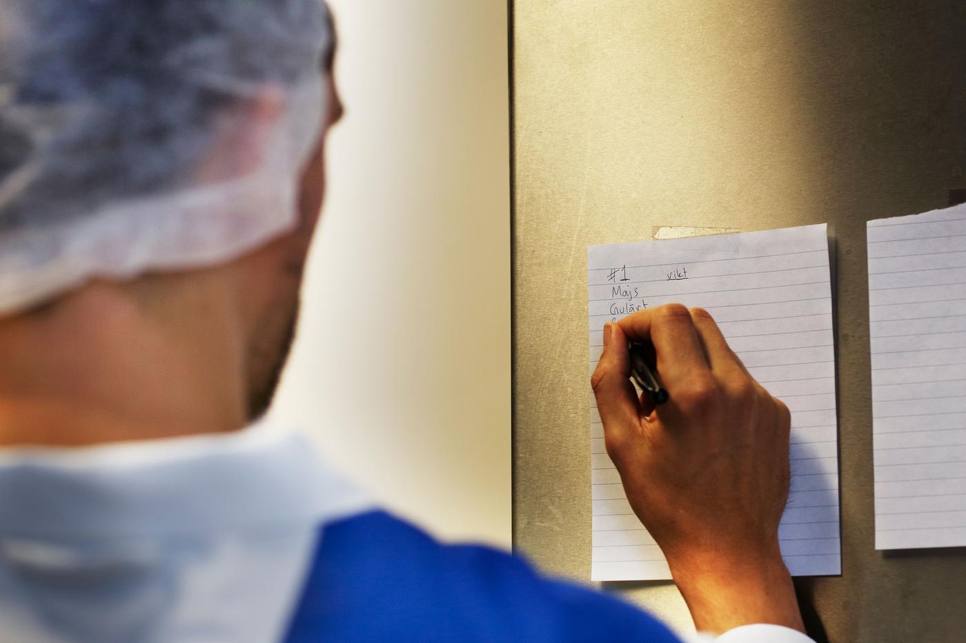 A person wearing a hairnet writes on a piece of notepaper on the wall.