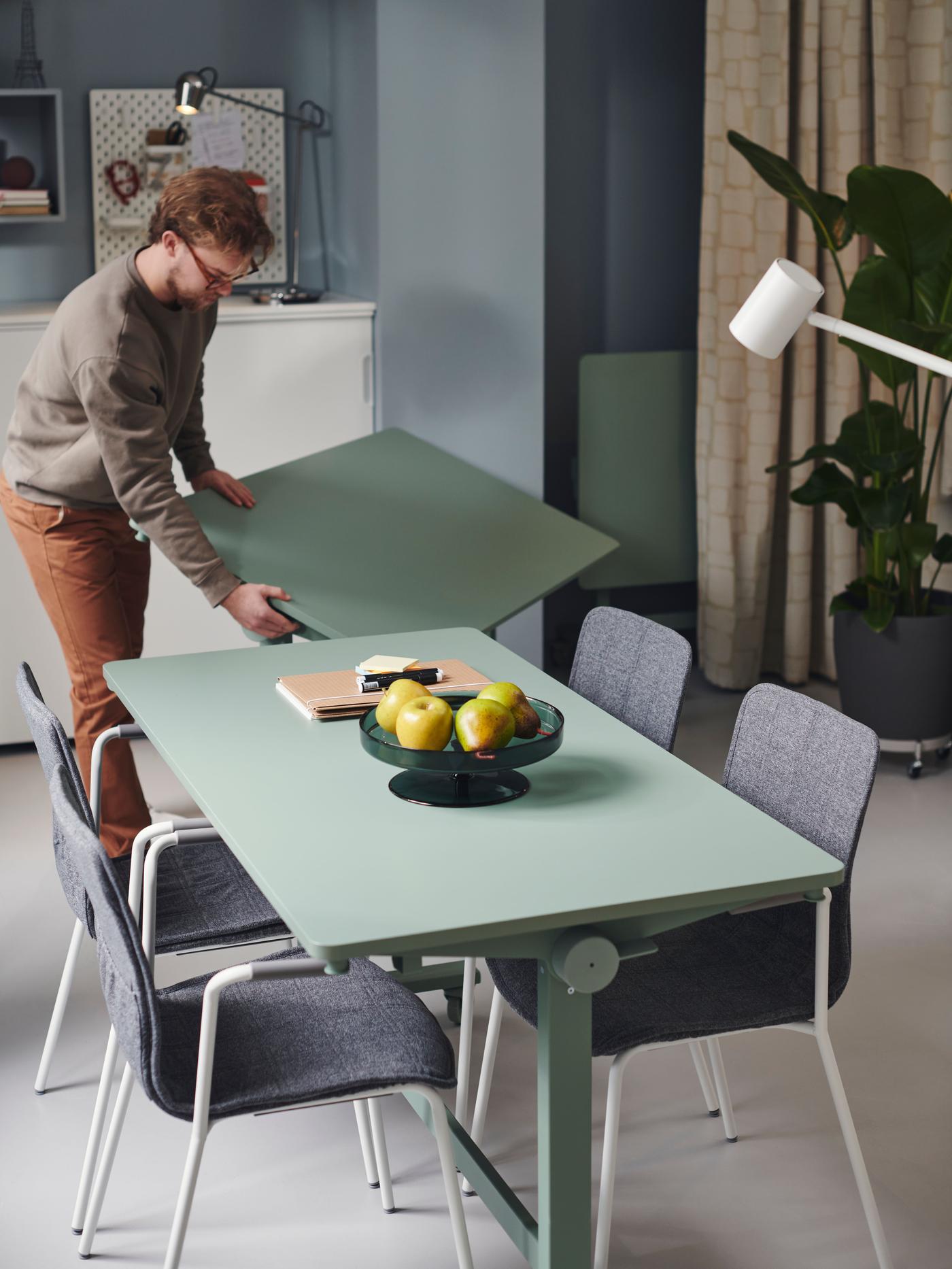 Green table with four grey chairs. On the table a basket of fruit and a man lifting another shorter green desk to place it next to the other.