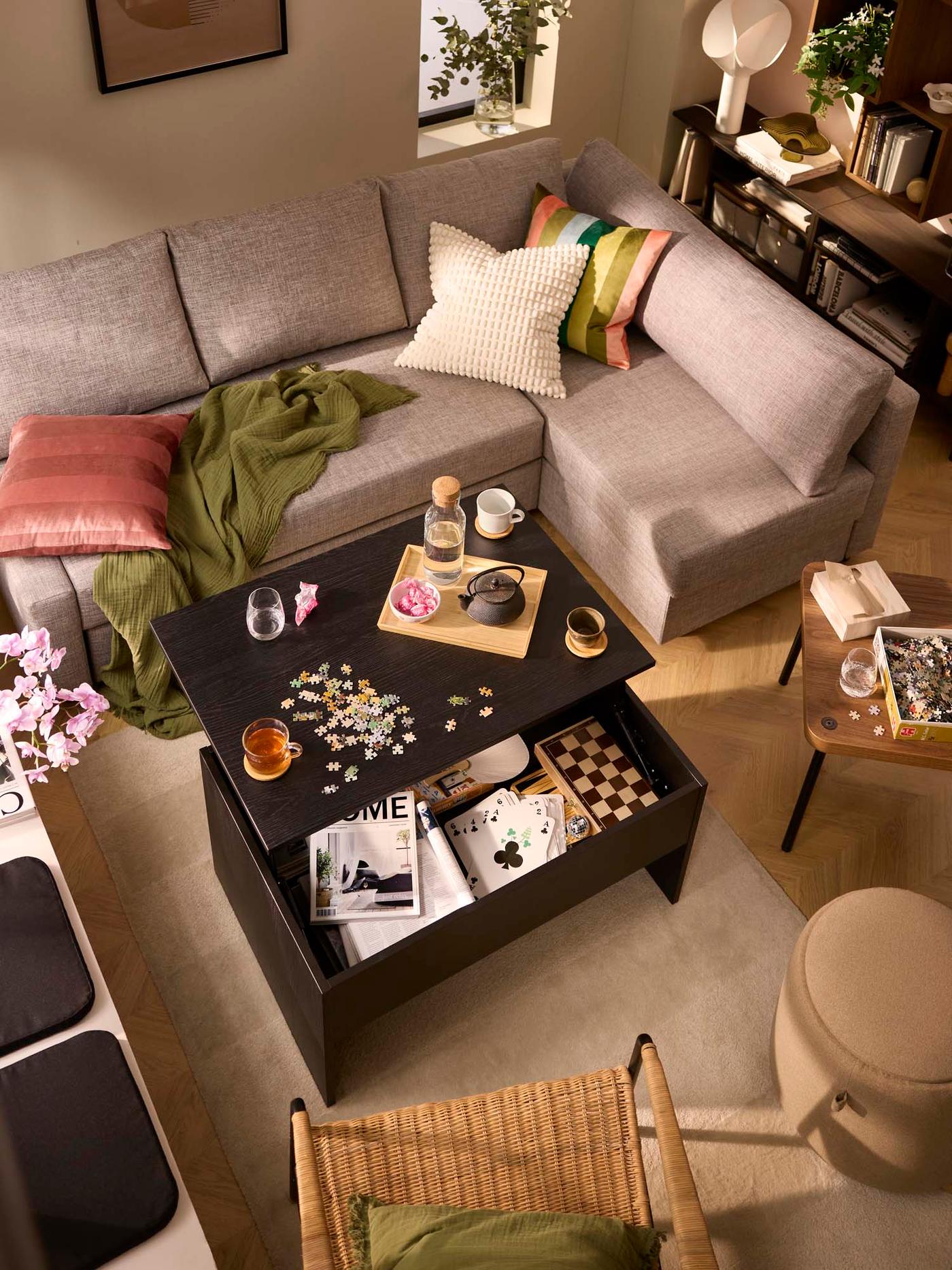 Top view of a cosy living room with a greyish tan sectional sofa, a black-brown coffee table in the middle set for board games, and storing decorative items.