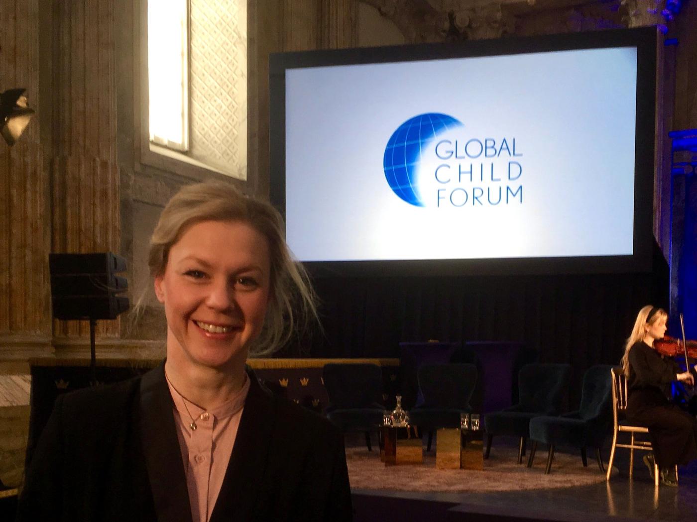 A person smiles at the camera with a presentation screen in the background reading "GLOBAL CHILD FORUM"