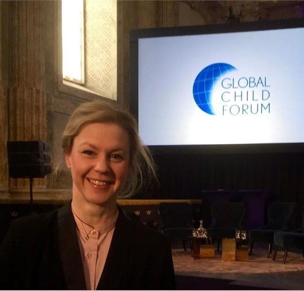 A person smiles at the camera with a presentation screen in the background reading "GLOBAL CHILD FORUM"