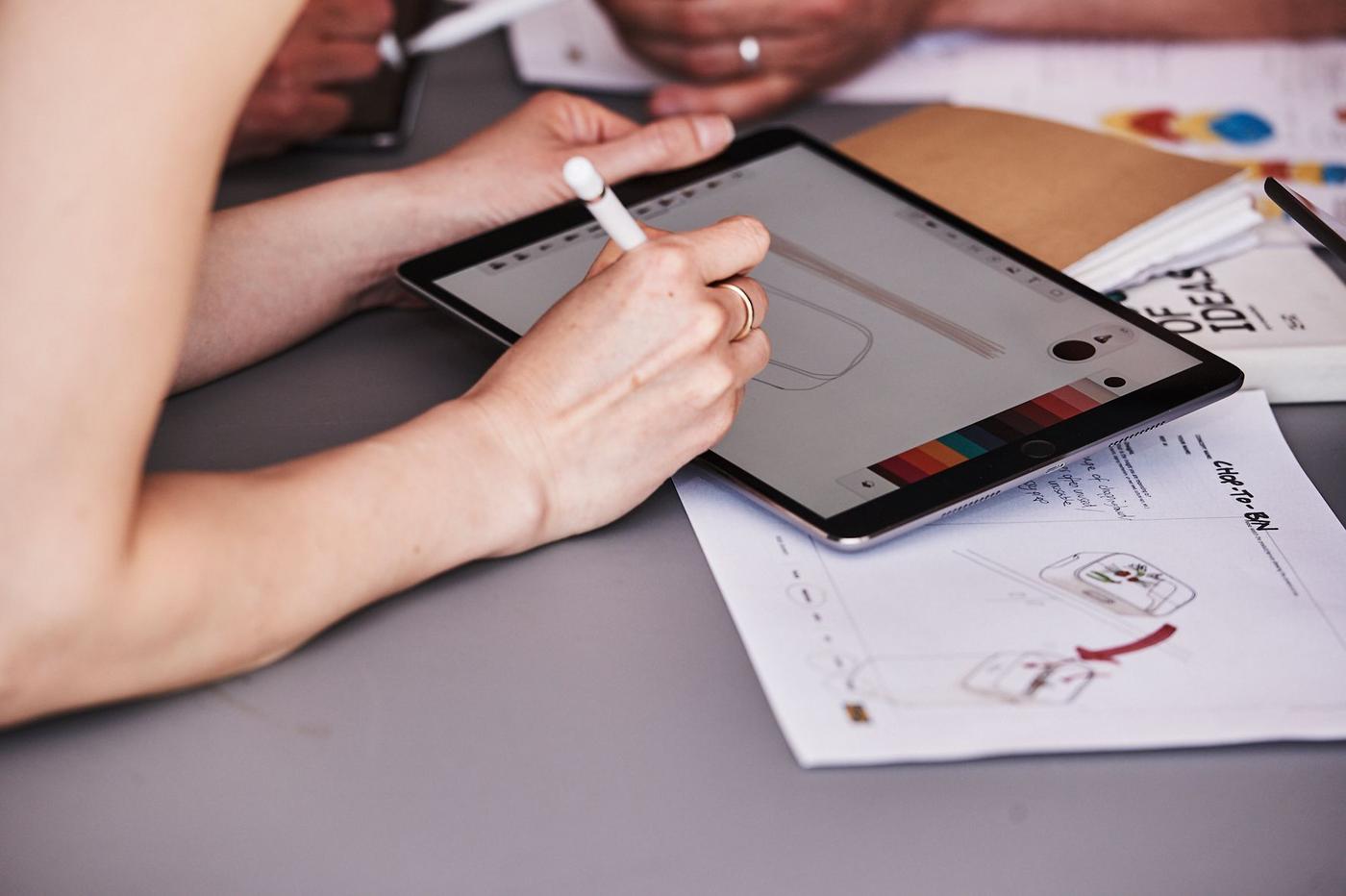 A tabletop covered in papers with a person sketching on a tablet.