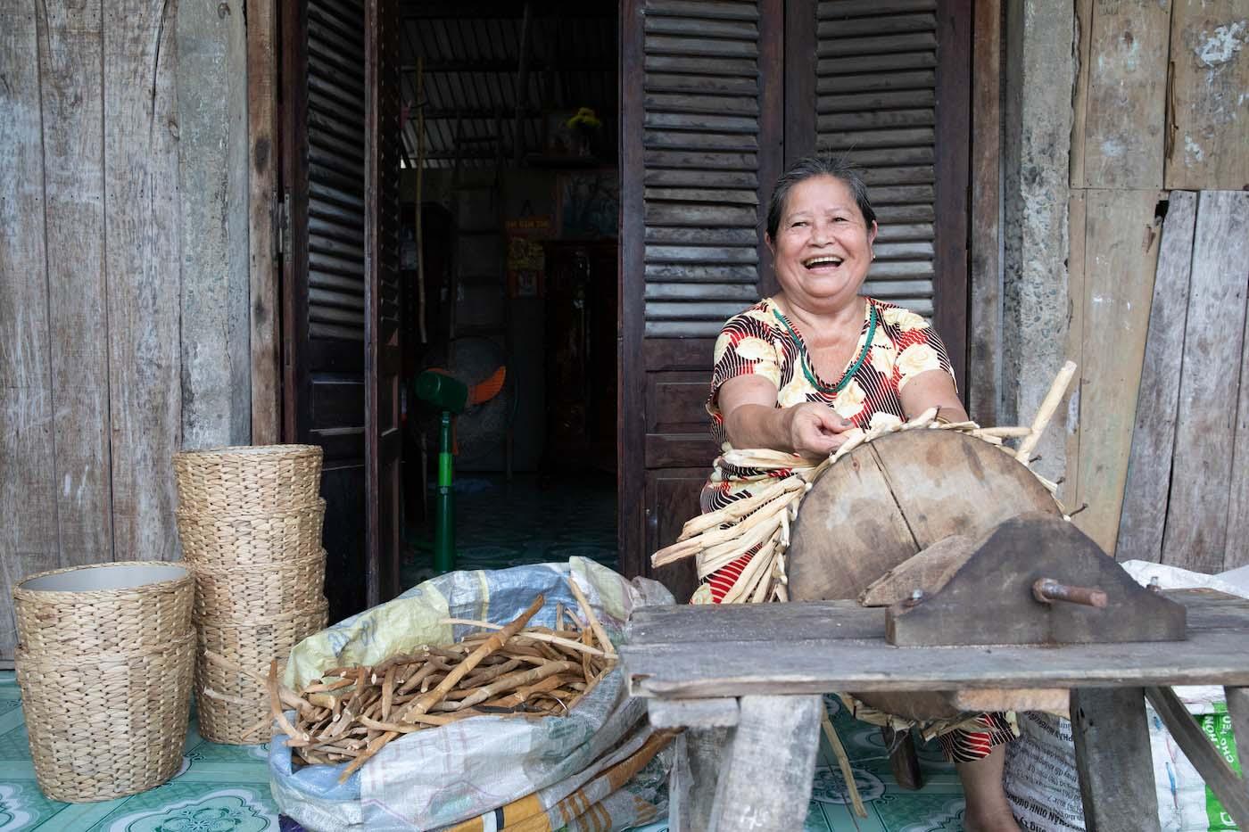 A person sits smiling as they weave a basket-like form from dried leaves.