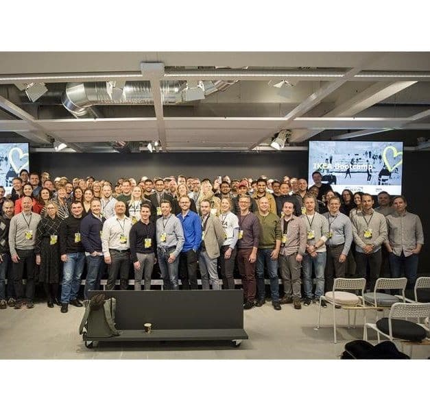 A large group of people stand in rows, looking at the camera. There is a TV screen behind them.