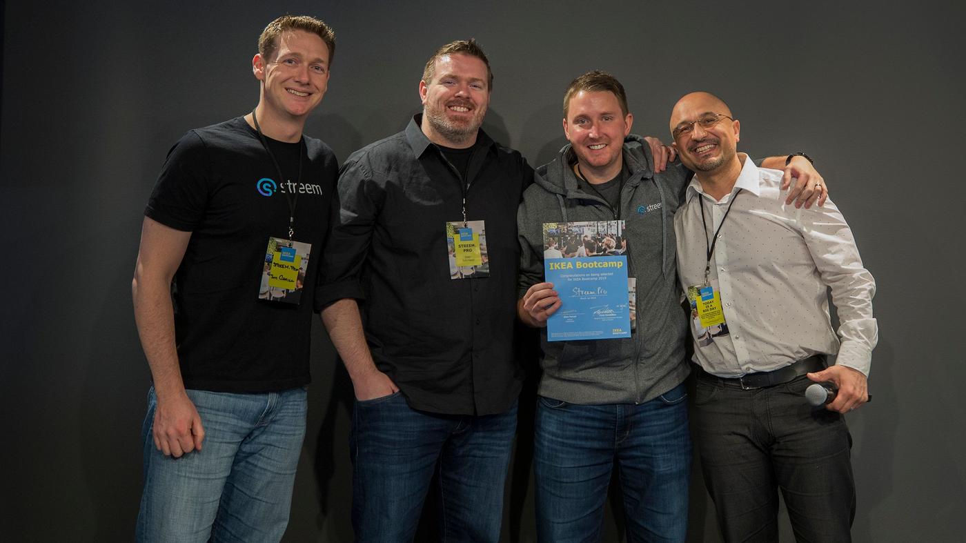 Four men smile at the camera. One is holding a microphone, the other holds a certificate.