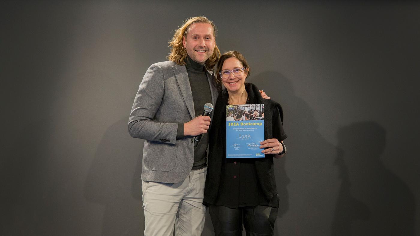 A man and woman smile at the camera. The man is holding a microphone and a certificate.