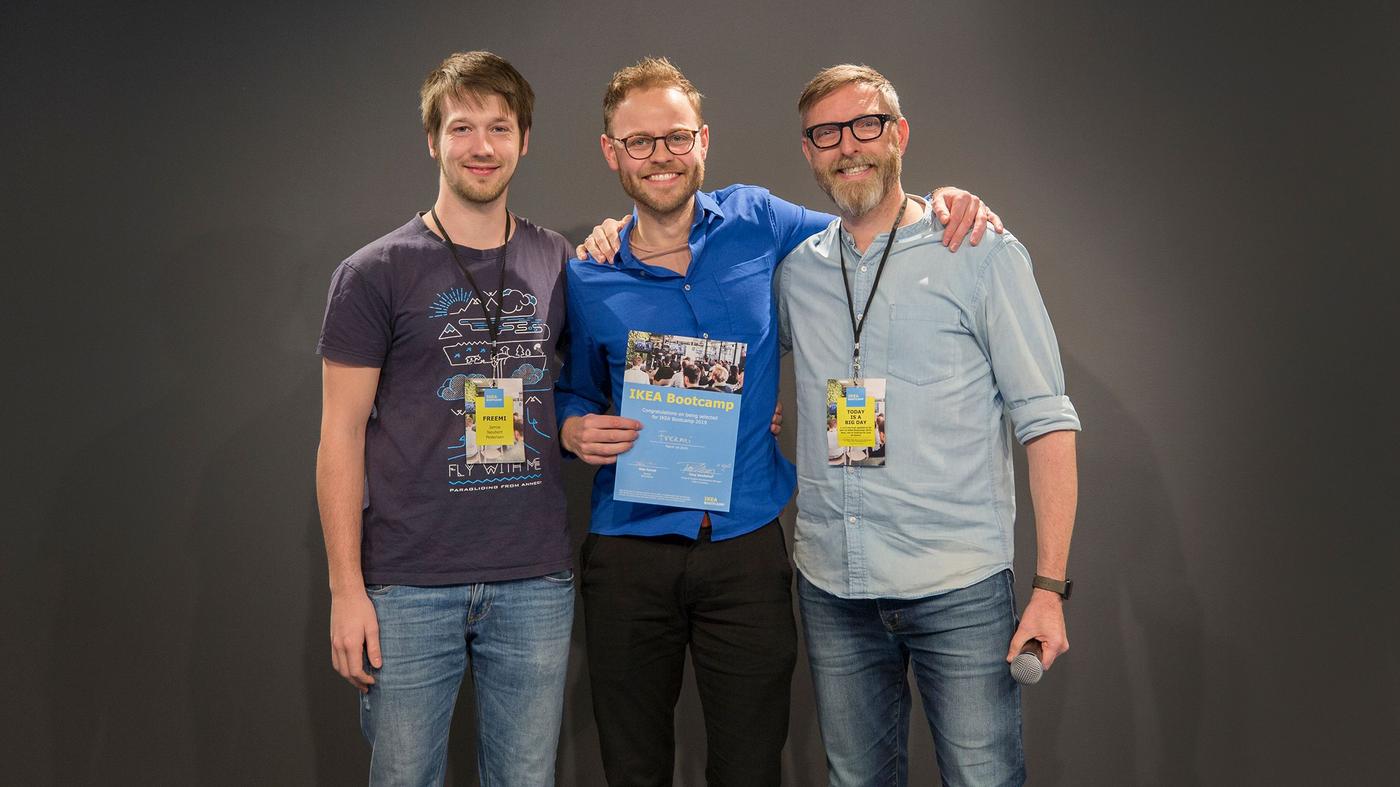Three men smile at the camera. One is holding a microphone, the other holds a certificate.