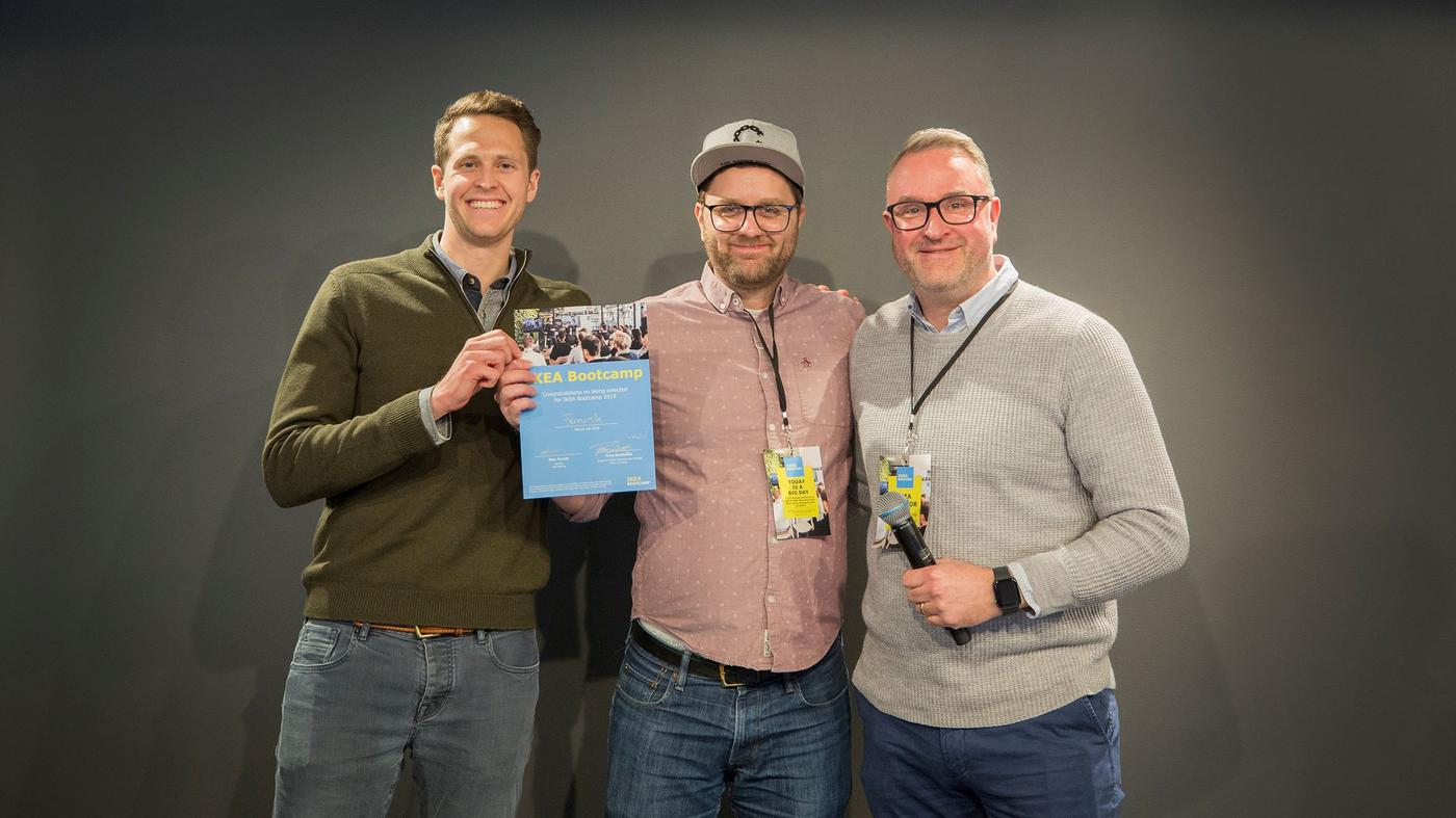 Three men smile at the camera. One is holding a microphone, the other holds a certificate.
