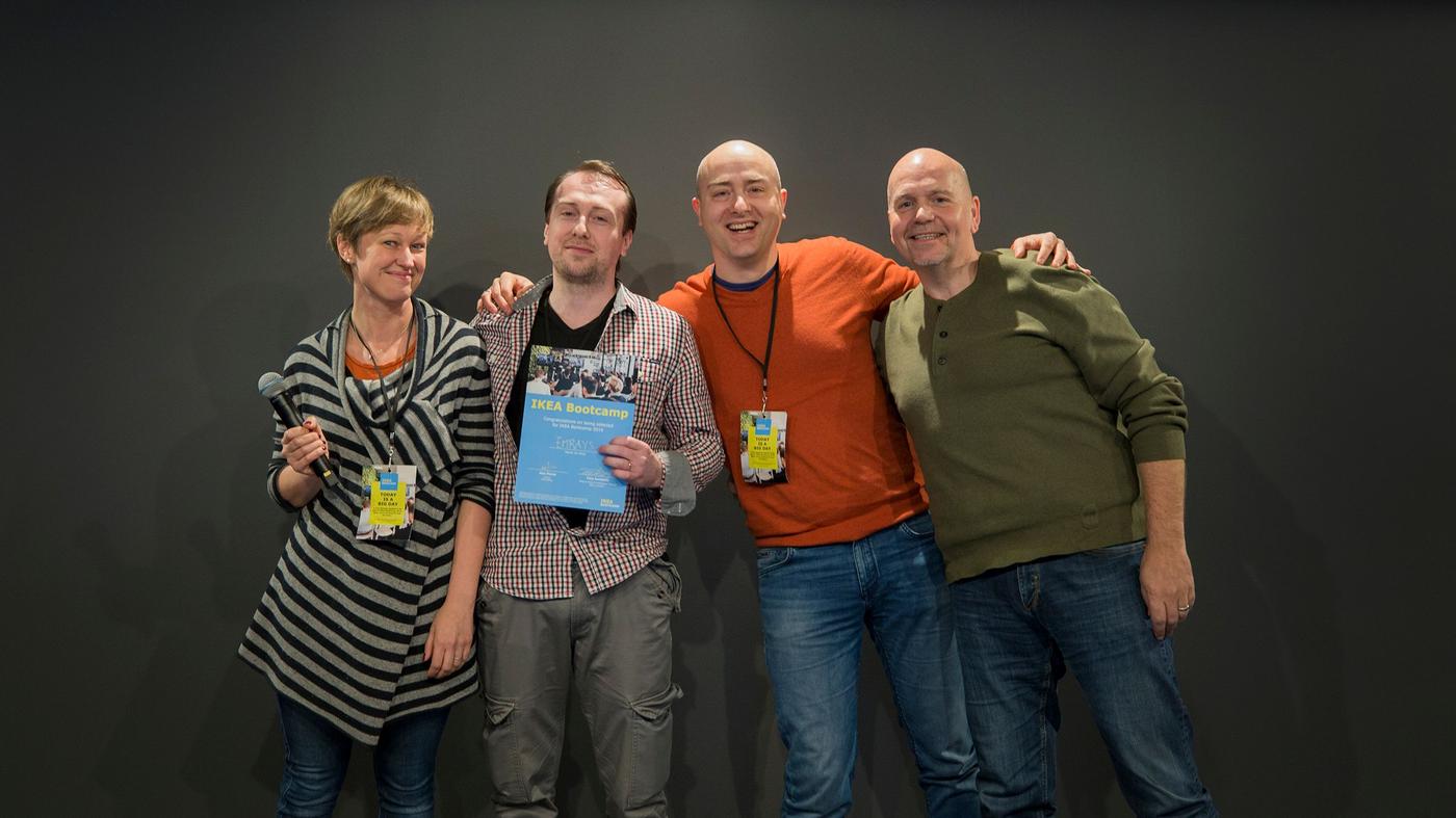 Two men and a woman smile at the camera. The woman is holding a microphone, a man holds a certificate.