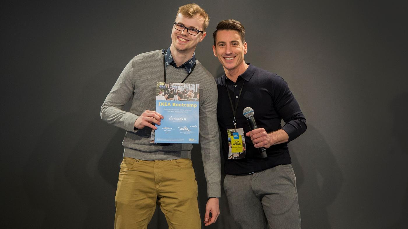 Two men smile at the camera. One is holding a microphone, the other holds a certificate.