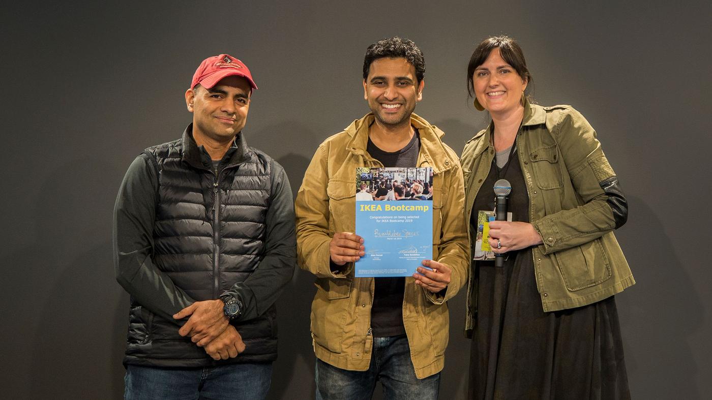 Two men and a woman smile at the camera. The woman is holding a microphone, the man holds a certificate.