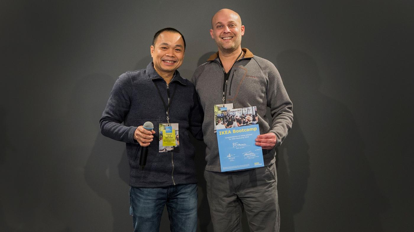 Two men smile at the camera. One is holding a microphone, the other holds a certificate.