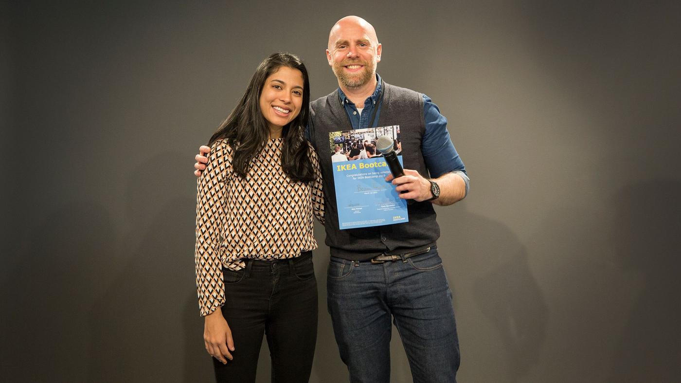 A man and woman smile at the camera. The man is holding a microphone and a certificate.