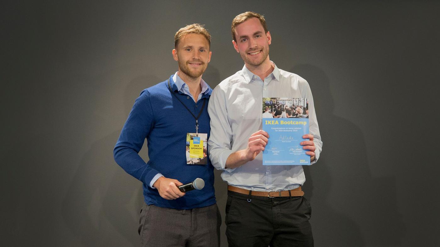 Two men smile at the camera. One is holding a microphone, the other holds a certificate.