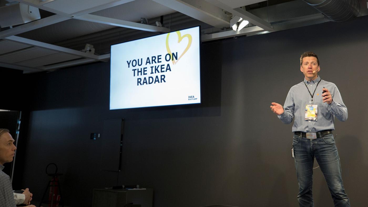 A man stands on a stage talking. There is a large screen near to him.