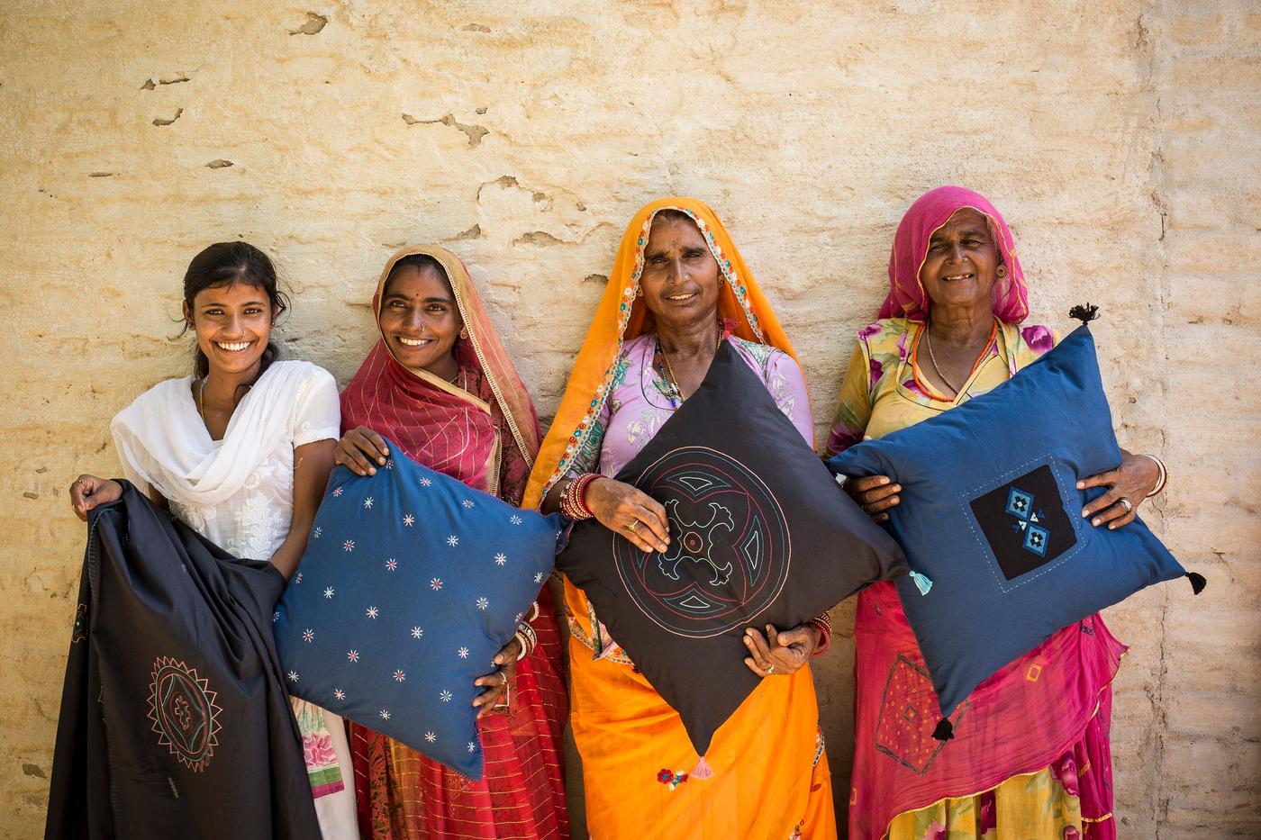 A row of four women wearing bright saris standing against a wall and each holding an embroidered cushion.