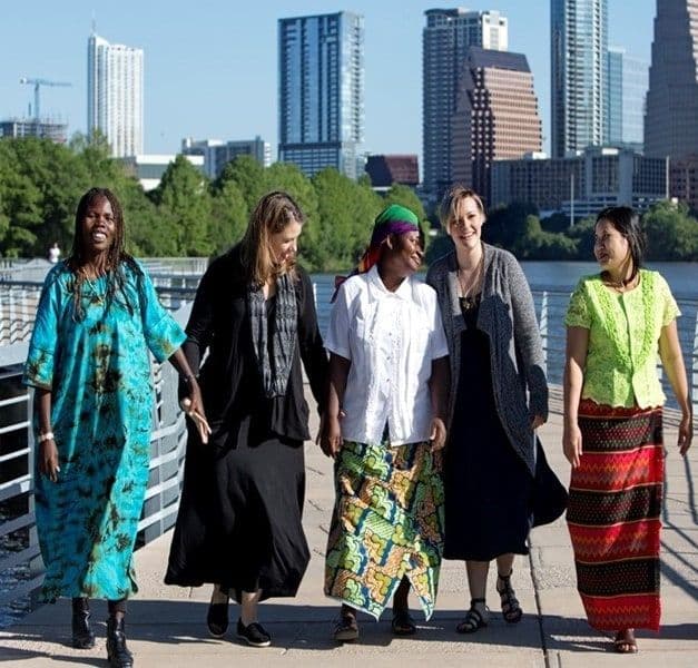 Five women stand in a row holding hands with a modern cityscape in the background.