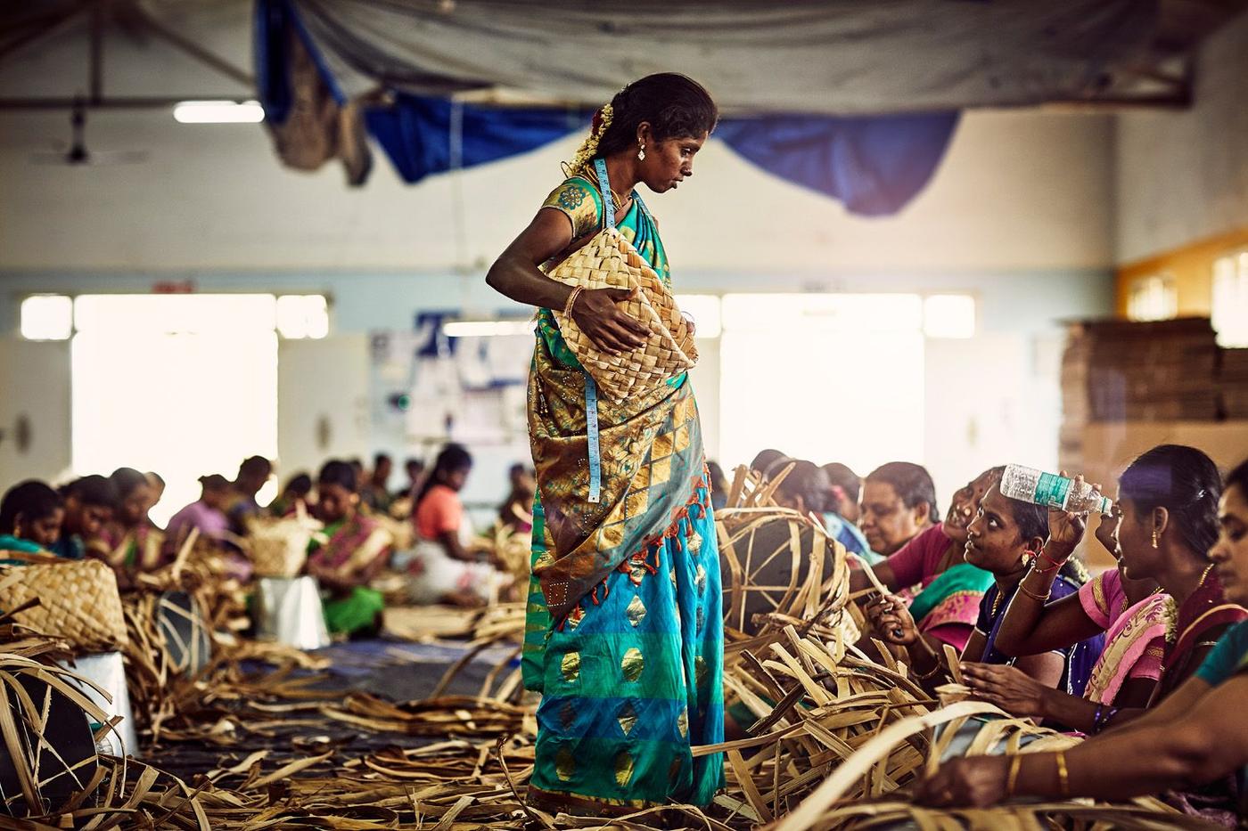 A large room of women weaving baskets from natural fibres. One stands holding a completed basket.