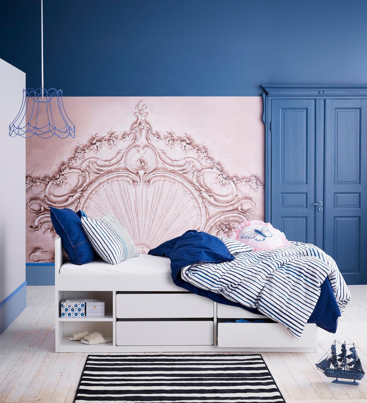 A white bed with built-in storage in a dark blue room with a pale pink accent wall.