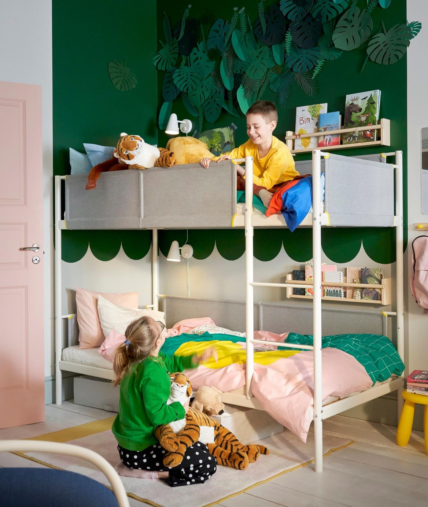 Two children in a bedroom with bunk bed and dark green walls. One sits on the floor looking at the other on the top bunk.